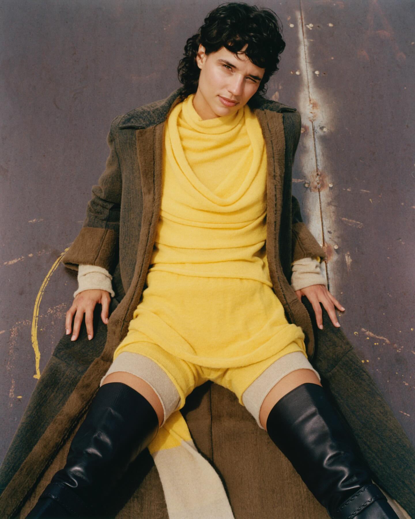 Fashion editorial shot of a young adult woman in a yellow outfit and black boots, posing in front of a weathered metal wall indoors.
