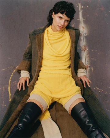 Fashion editorial shot of a young adult woman in a yellow outfit and black boots, posing in front of a weathered metal wall indoors.
