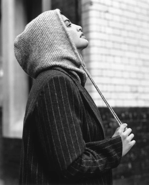 Monochrome portrait of a woman in pinstripe blazer and knitted hood looking up, standing in an urban alleyway with brick walls.