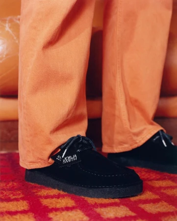 Close-up studio shot of black suede Clarks x Jorja Smith shoes on orange patterned rug, featuring orange trousers and couch.