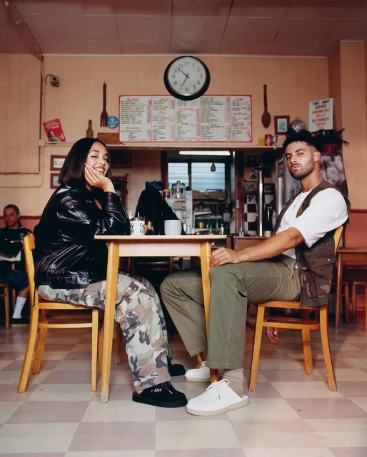 A young couple is sitting at a table in a vintage-style diner, engaged in conversation. The scene is bathed in warm, natural light.