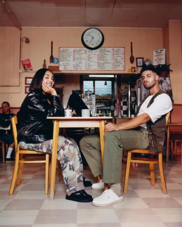 A young couple is sitting at a table in a vintage-style diner, engaged in conversation. The scene is bathed in warm, natural light.