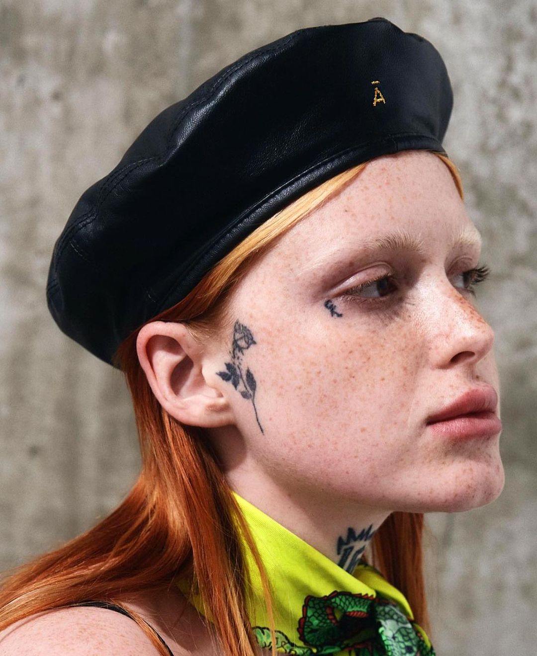Close-up of a red-haired adult female in a black leather beret with a gold emblem, tattoos on her face and neck, and light skin.