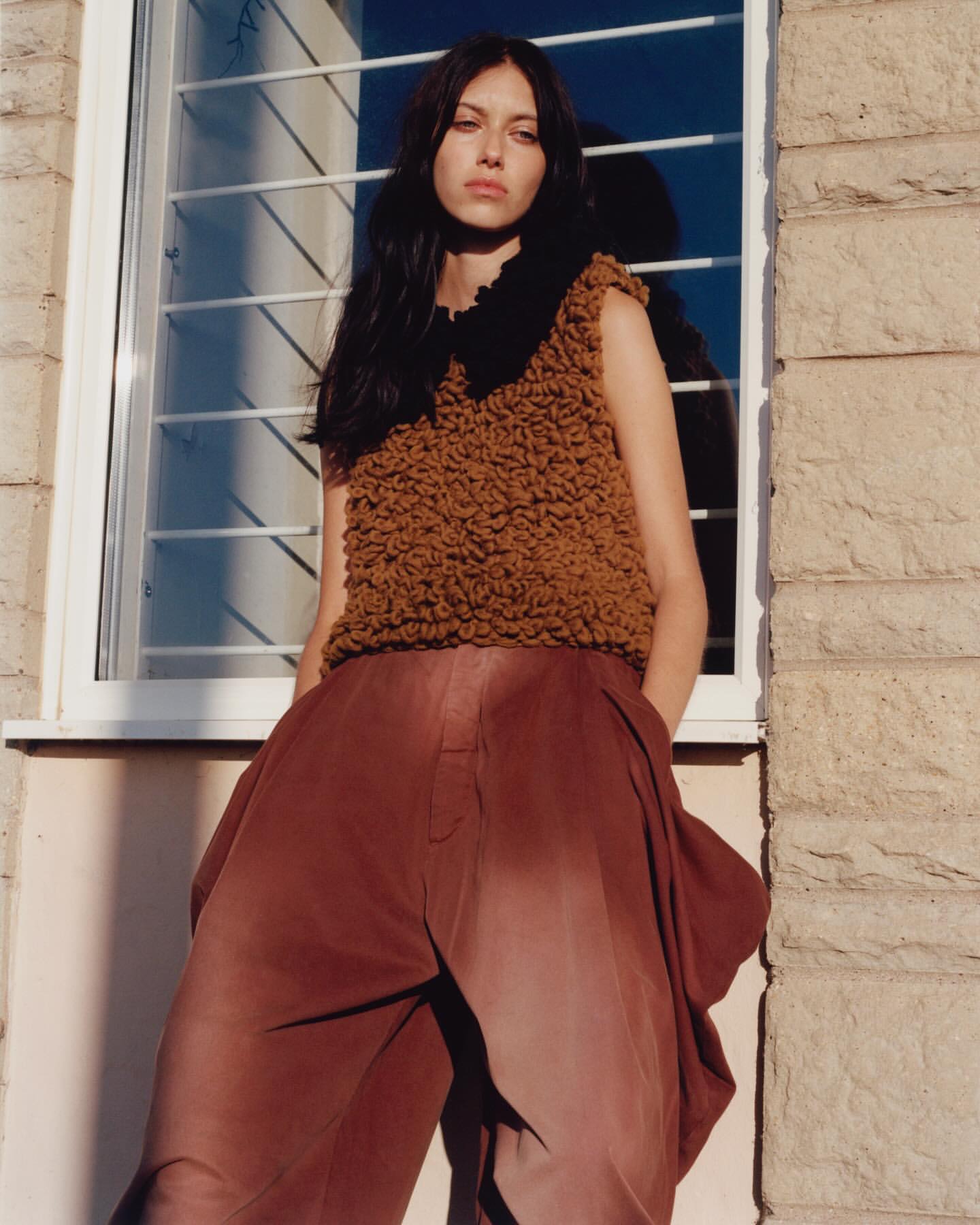 A young adult woman with dark hair poses in a brown, textured sleeveless top and flowing red-brown trousers, standing indoors near a window.
