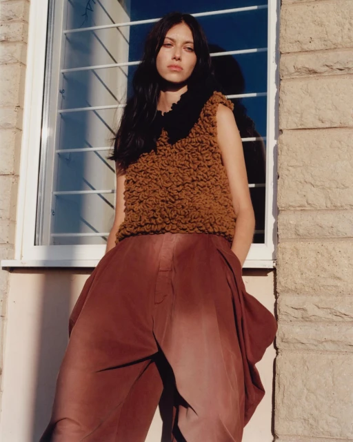 A young adult woman with dark hair poses in a brown, textured sleeveless top and flowing red-brown trousers, standing indoors near a window.