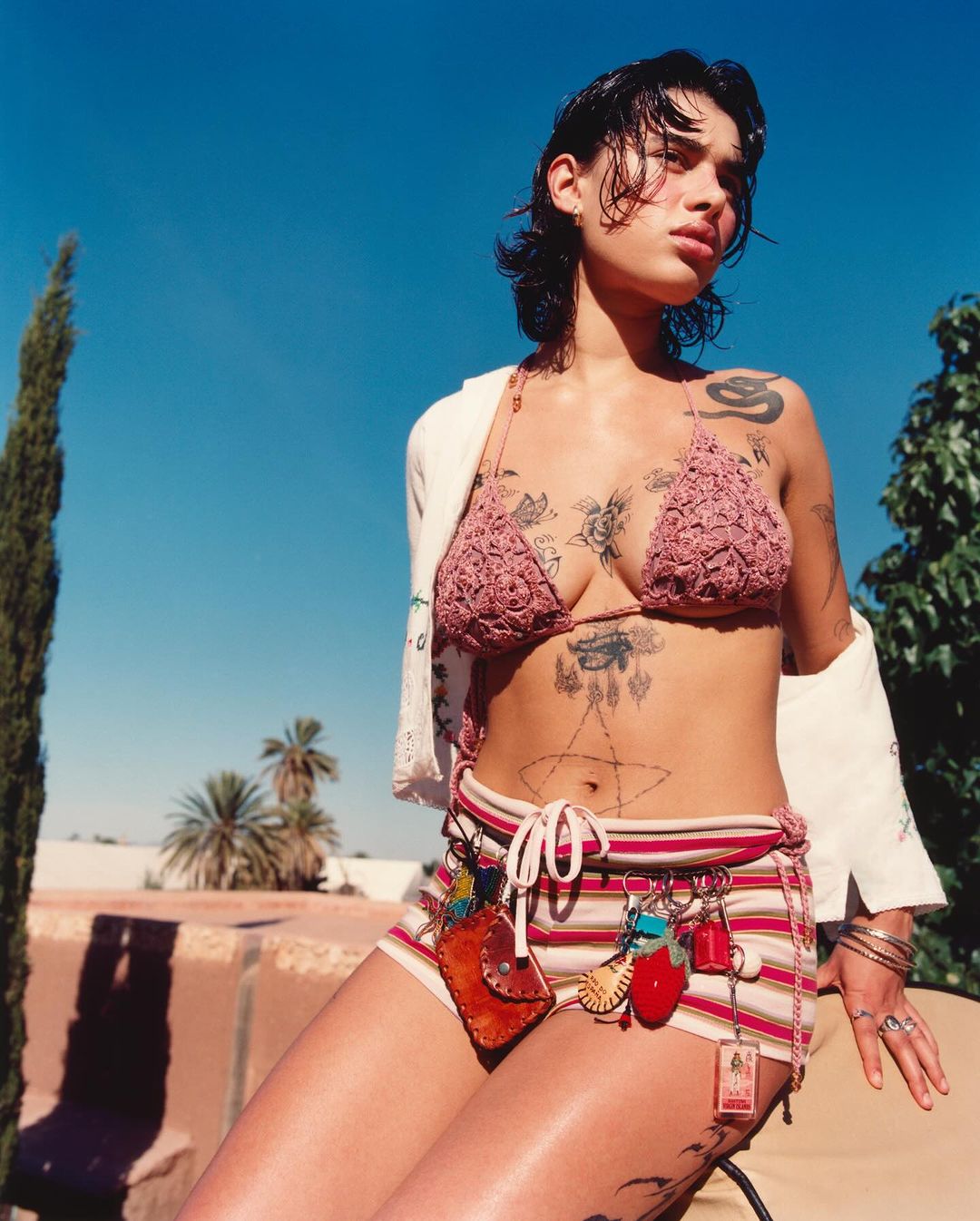 Young woman poses outdoors in a crochet bikini top and striped shorts with charms, set against a blue sky and green foliage backdrop.