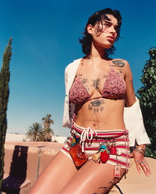 Young woman poses outdoors in a crochet bikini top and striped shorts with charms, set against a blue sky and green foliage backdrop.