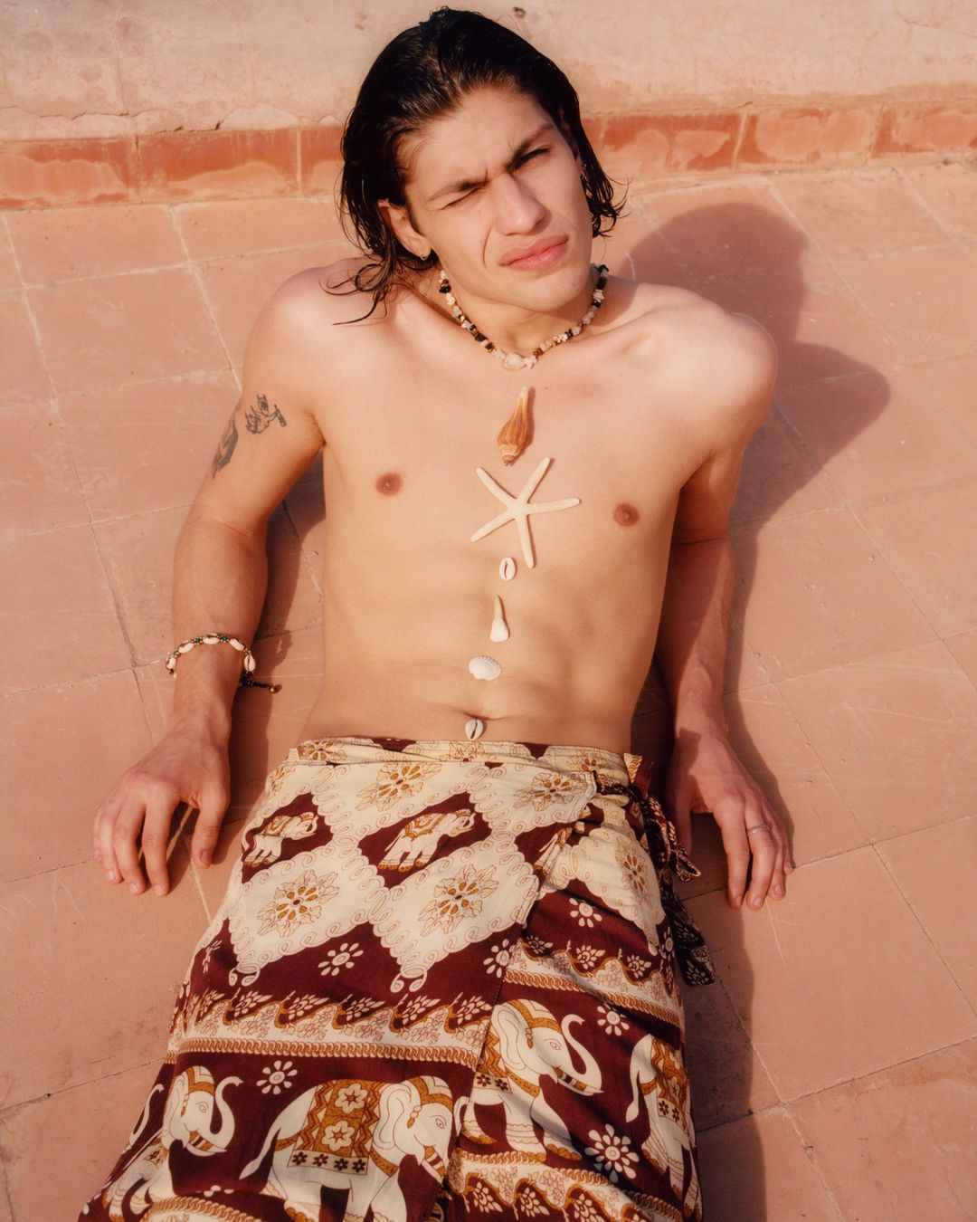 A shirtless man lies on a tile surface wearing a patterned sarong with shells and a starfish arranged on his torso, shot outdoors.
