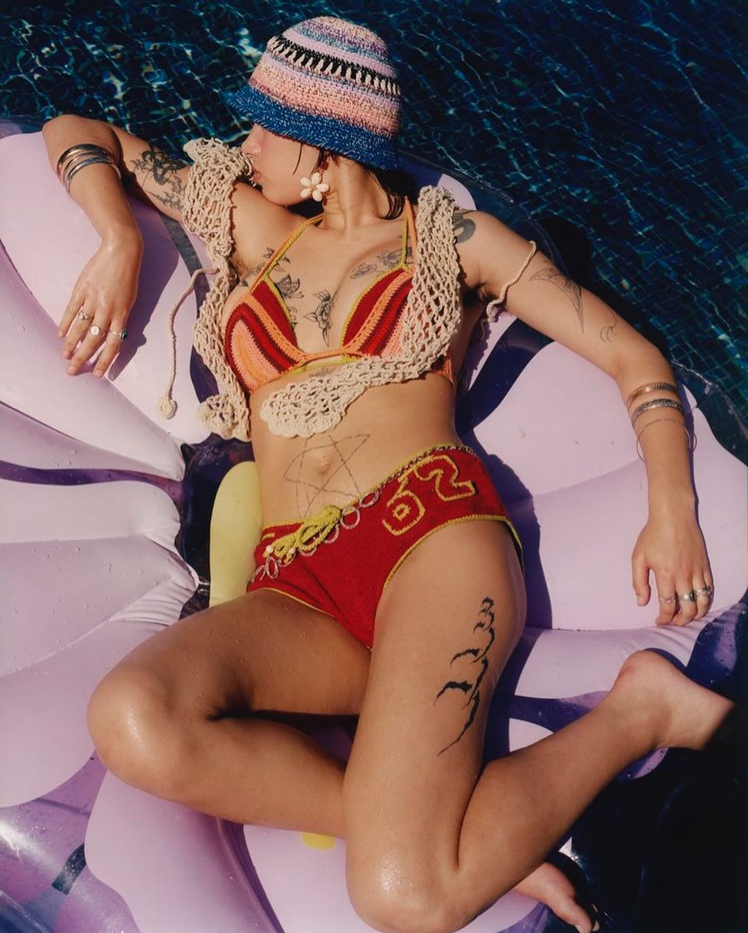 A woman with tattoos lounges on a flower-shaped pool float, wearing a knitted bikini and bucket hat, enjoying a sunny day in the water.