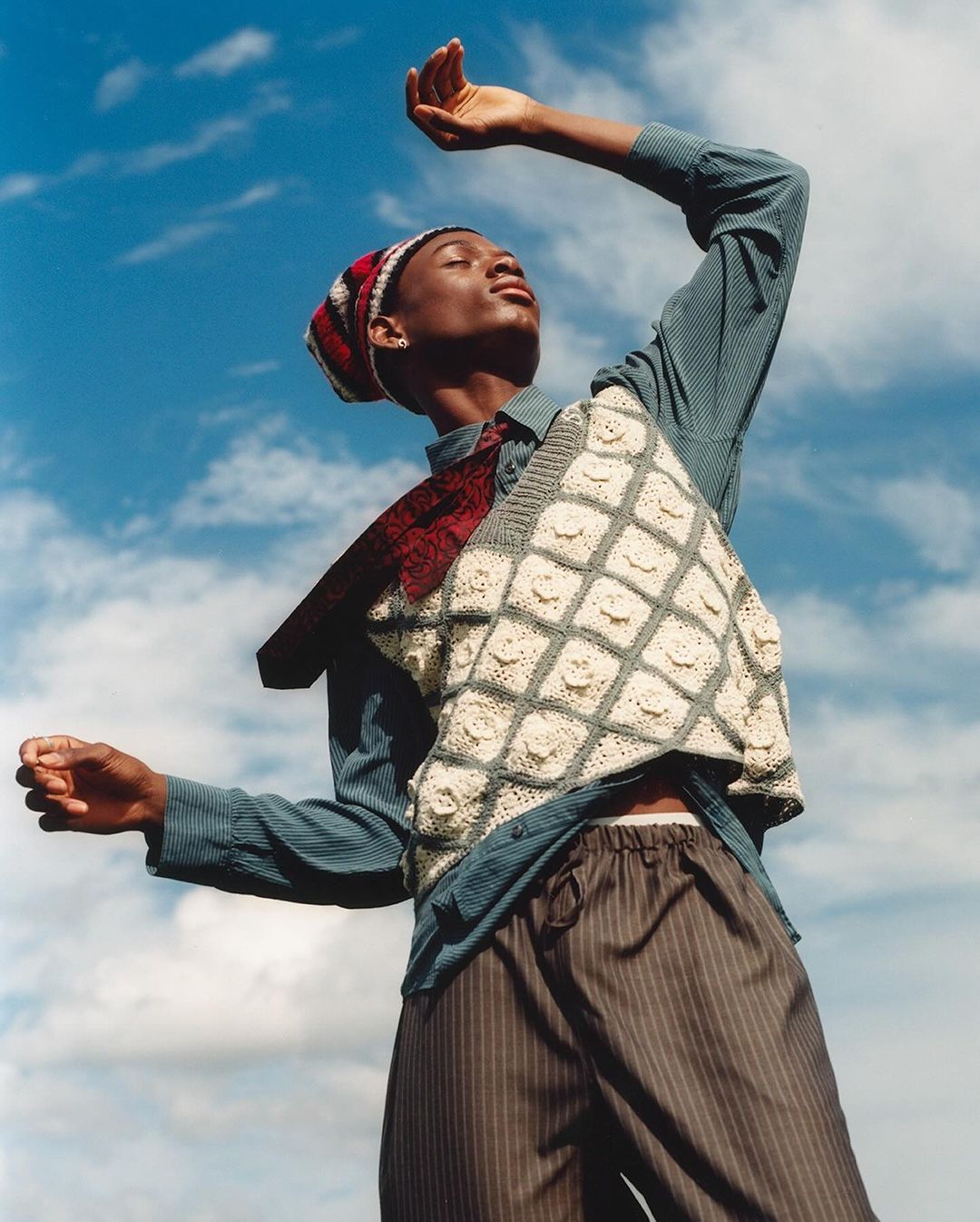 Young adult person poses against a blue sky wearing a casual shirt, striped pants, sweater vest, and beanie with raised arms.
