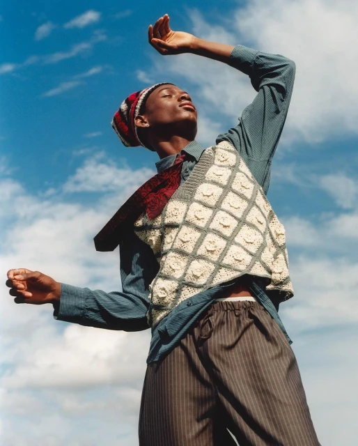 Young adult person poses against a blue sky wearing a casual shirt, striped pants, sweater vest, and beanie with raised arms.