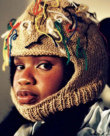 Eye-level portrait of an adult female with dark skin and glossy lips wearing a tan knit balaclava-style hat with colorful yarn strands.