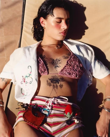 Young woman with dark hair and chest tattoos resting outdoors in a crocheted bikini top and striped shorts photographed by Kaj Jefferies.
