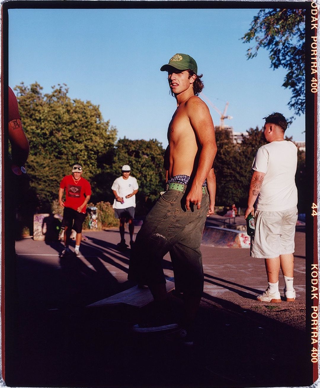 A shirtless adult male wears a baseball cap while standing on a skateboard in an outdoor skatepark on a sunny day, surrounded by other people.