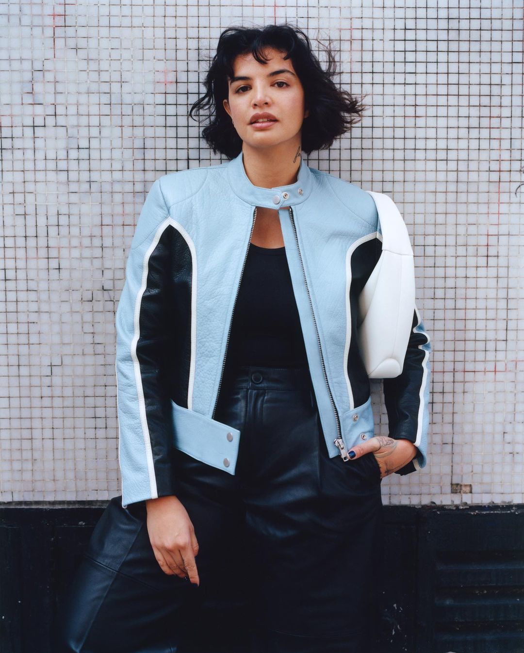 Woman with dark short hair wearing a light blue and black leather jacket against a tiled wall by Kaj Jefferies