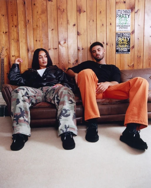 Two people sit together on a brown leather couch in a casual setting with a wood-paneled wall. Man in orange pants, woman in camo.