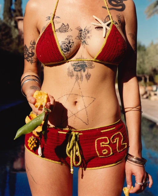 Woman in red crocheted bikini and shorts holds a cut orange outdoors with blue pool tiles, tattoos, and yellow details.