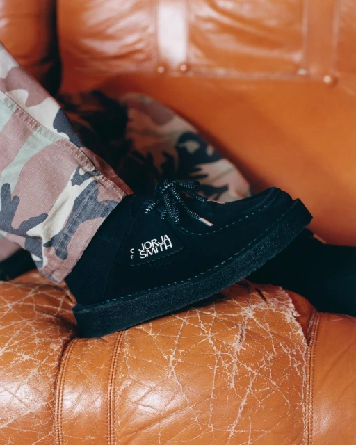 Black Clarks shoe with Jorja Smith branding, worn with camouflage pants, rests on a worn orange leather couch.