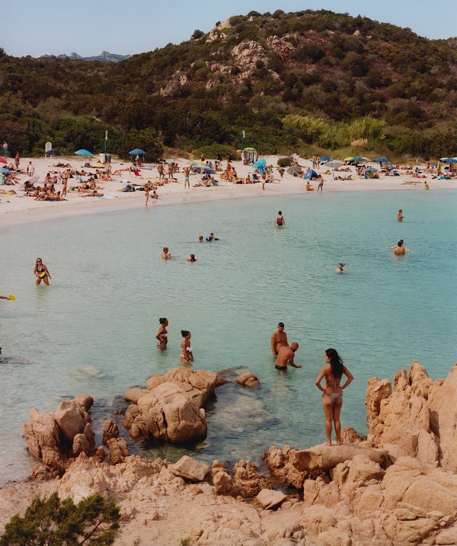 Coastal beach scene with people swimming, sunbathing, and relaxing on the sandy shore, backed by a hill covered with trees and vegetation.