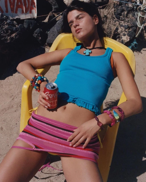 Young woman in a blue tank top and pink striped skirt relaxes in a yellow chair on a beach with a Coca-Cola can in hand.
