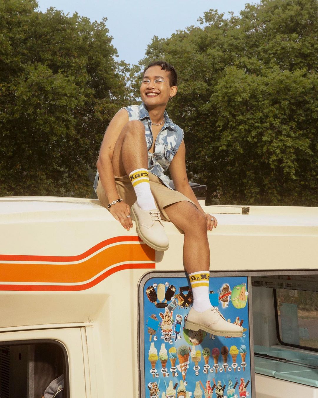 Adult male sitting on top of a car wearing shorts, a denim vest, and Dr. Martens shoes with trees in the background.
