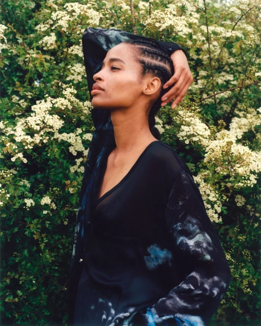 Portrait of adult Black woman with cornrows, looking up in an outdoor setting with green foliage and white flowers behind her.