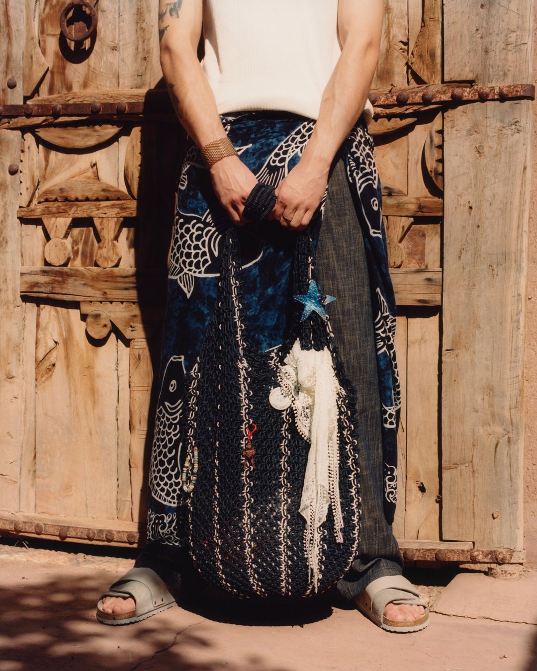 Person in sarong and sandals holds a mesh bag in front of an ornate wooden door in an outdoor setting, casting a warm shadow.