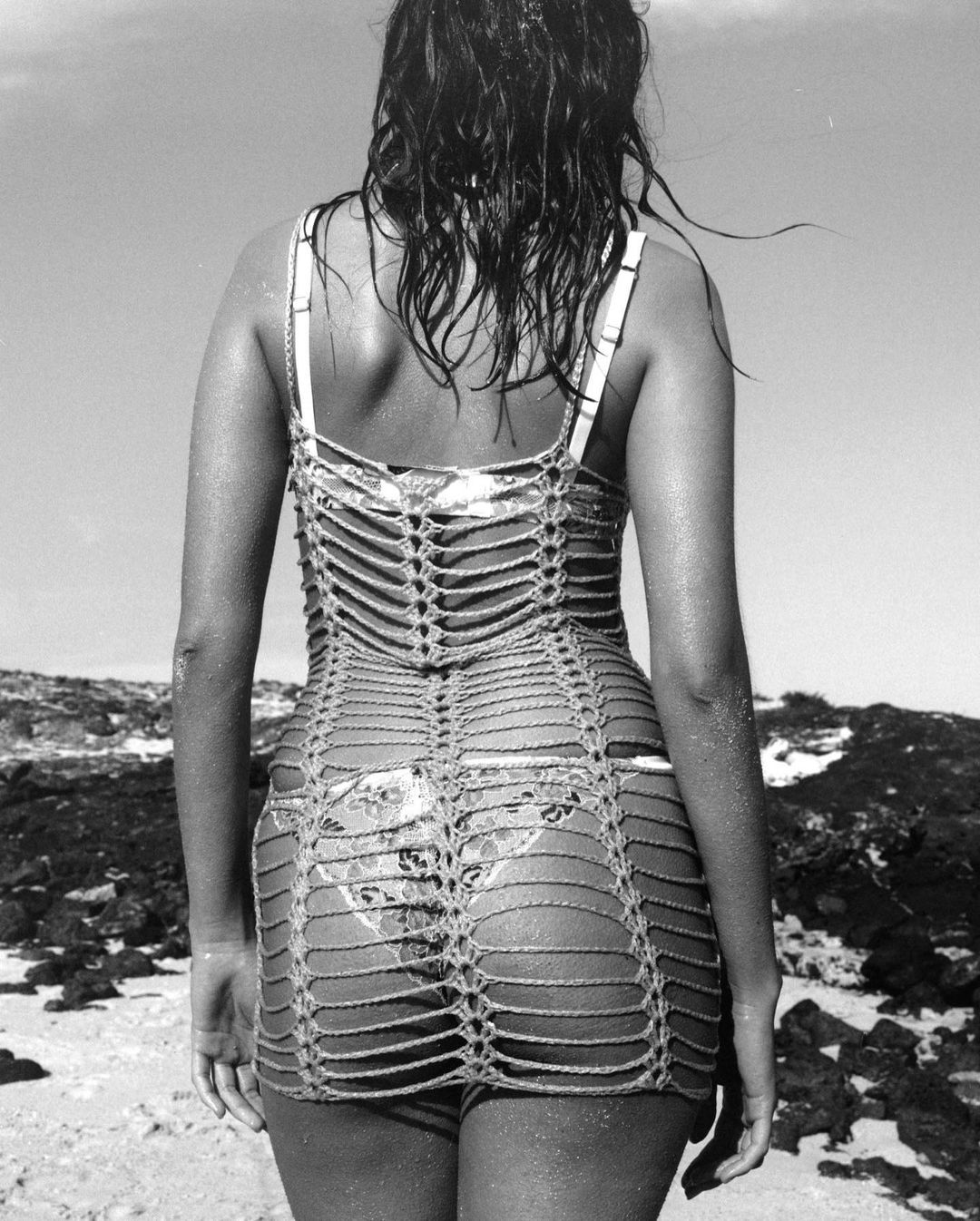 Black and white portrait of an adult woman with wet hair, wearing a cover-up dress on a beach with rocks on a sunny day.
