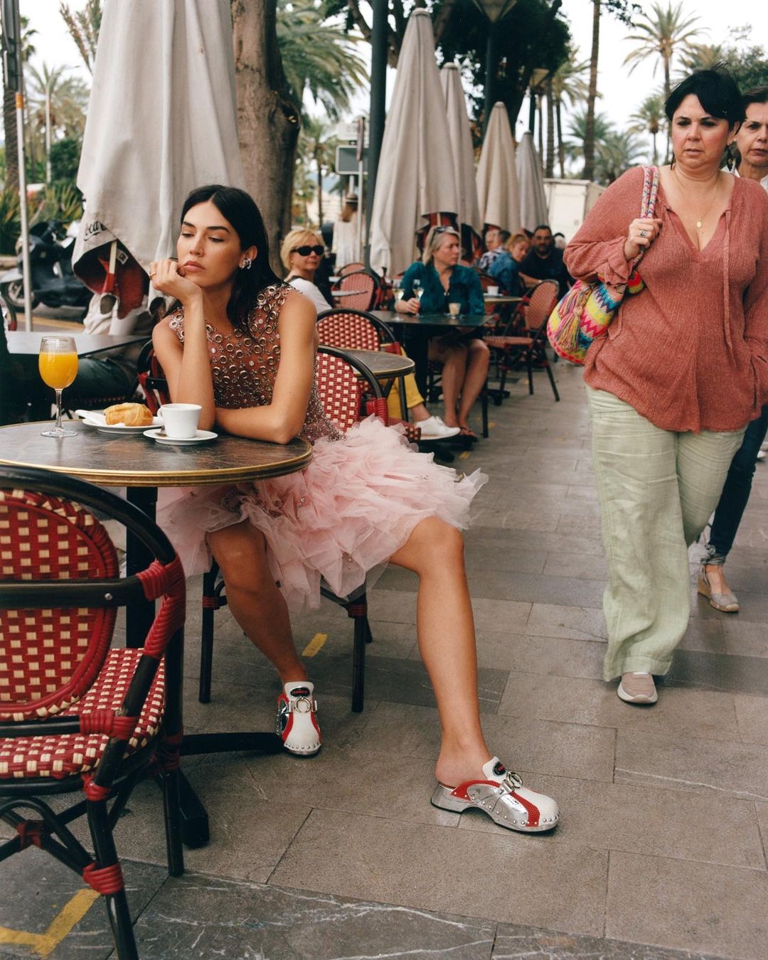 A young adult woman sits at a cafe table with a drink and pastry, wearing a dress and stylish shoes on an outdoor city street.