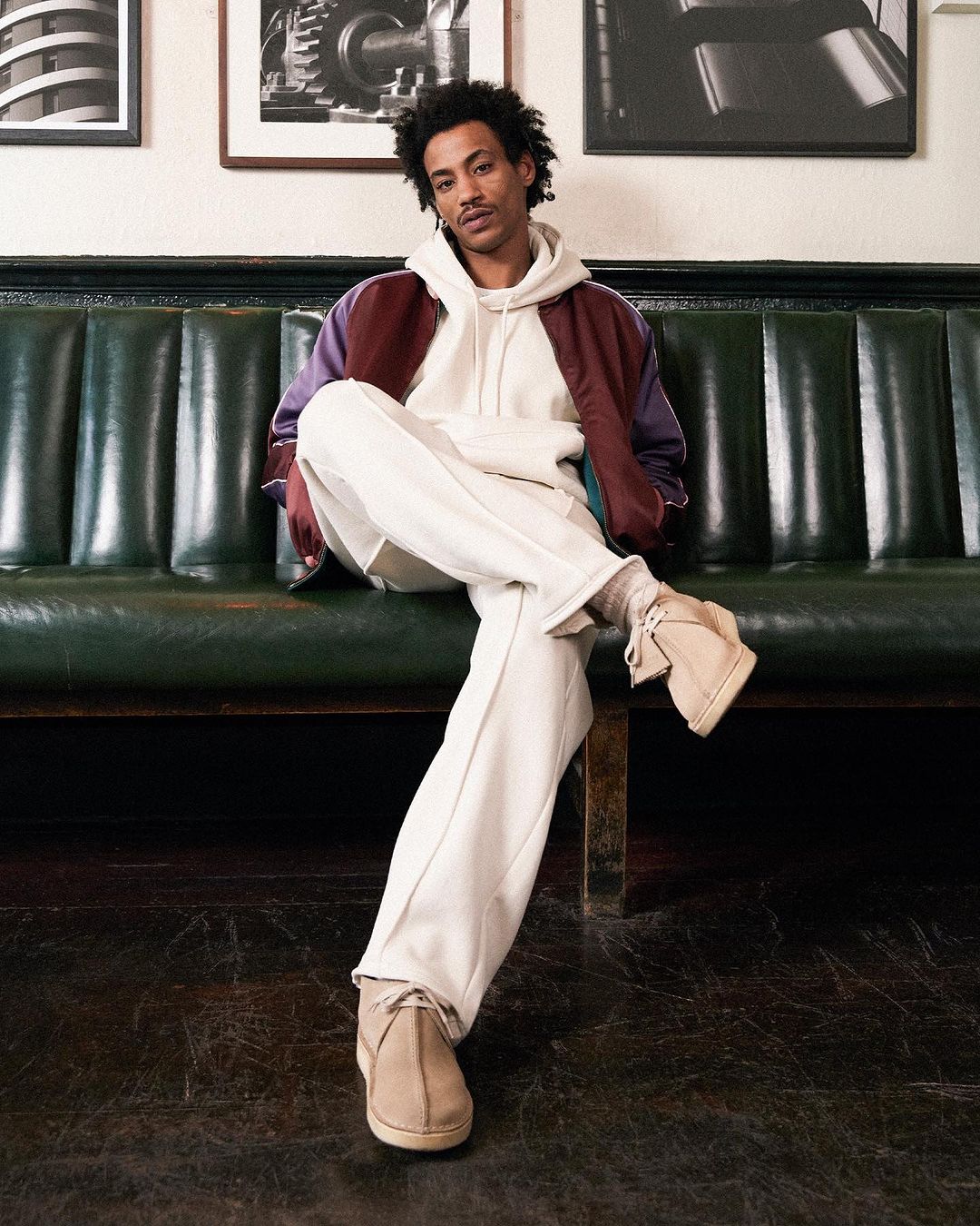 A young adult male sits on a green leather bench in casual wear. He wears a hoodie, jacket, cream pants, and desert boots, indoors.
