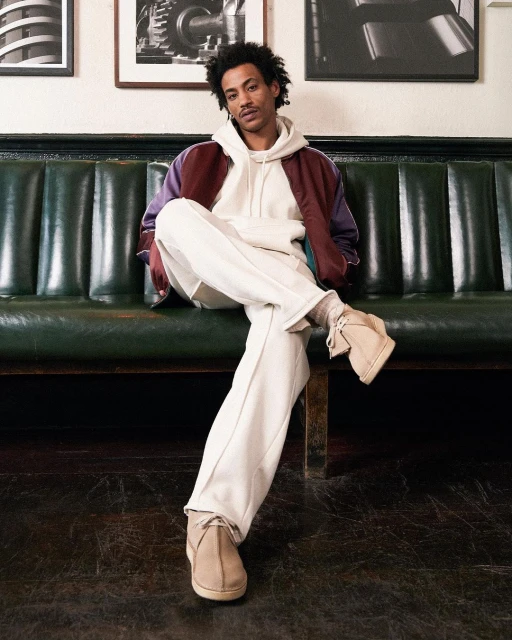 A young adult male sits on a green leather bench in casual wear. He wears a hoodie, jacket, cream pants, and desert boots, indoors.
