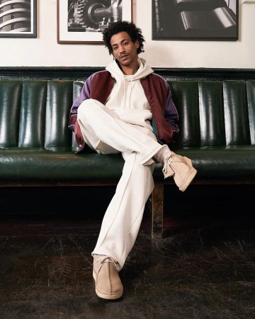 A young adult male sits on a green leather bench in casual wear. He wears a hoodie, jacket, cream pants, and desert boots, indoors.