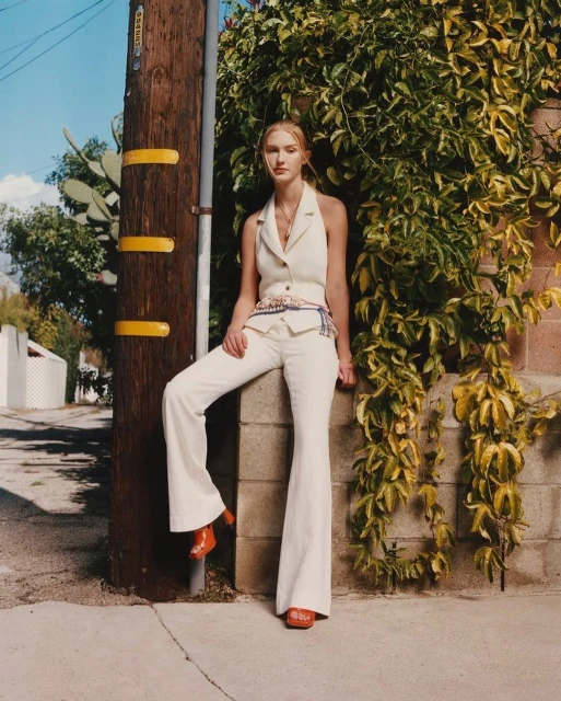 A woman in a cream halter top and bell-bottom pants leans against a telephone pole next to a brick wall covered in ivy outdoors.