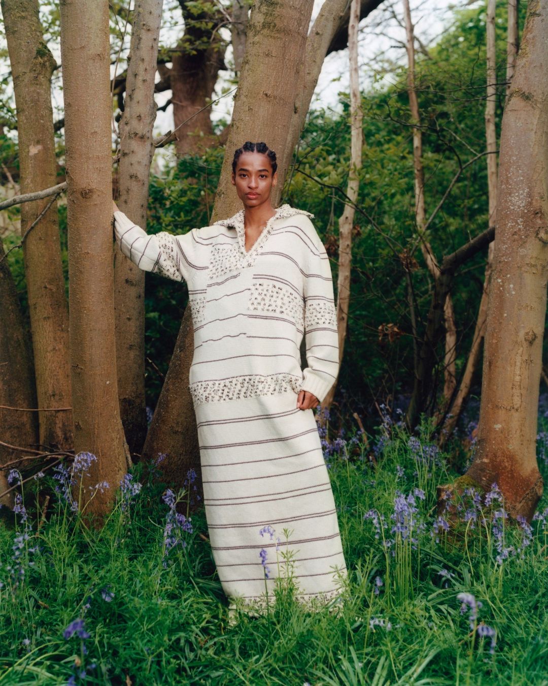 A woman in a patterned white dress poses amongst trees in a forest of bluebell flowers in an outdoor fashion shot.