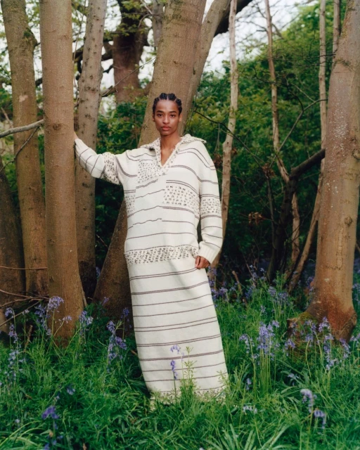 A woman in a patterned white dress poses amongst trees in a forest of bluebell flowers in an outdoor fashion shot.