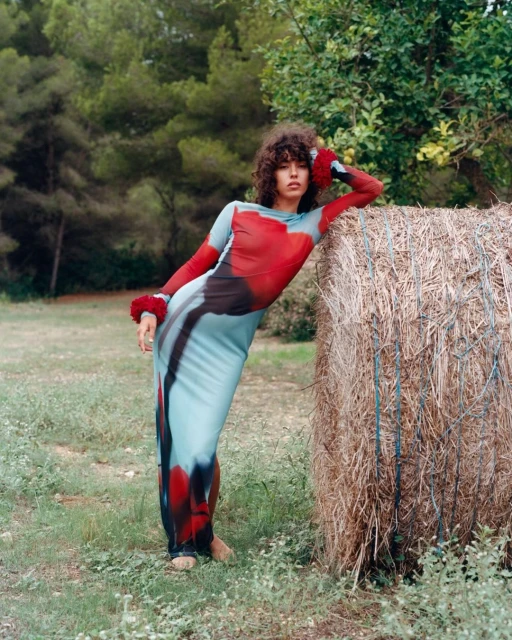 An adult woman with curly hair poses in a long patterned dress next to a hay bale in a grassy field with trees in the background.