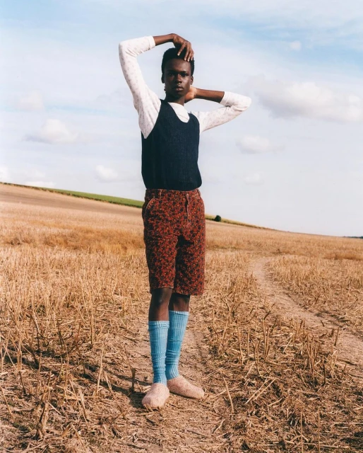 A stylish adult stands in a field, wearing a sweater vest, printed shorts, and knee-high socks. Rural setting with blue sky.