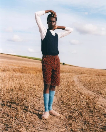 A stylish adult stands in a field, wearing a sweater vest, printed shorts, and knee-high socks. Rural setting with blue sky.