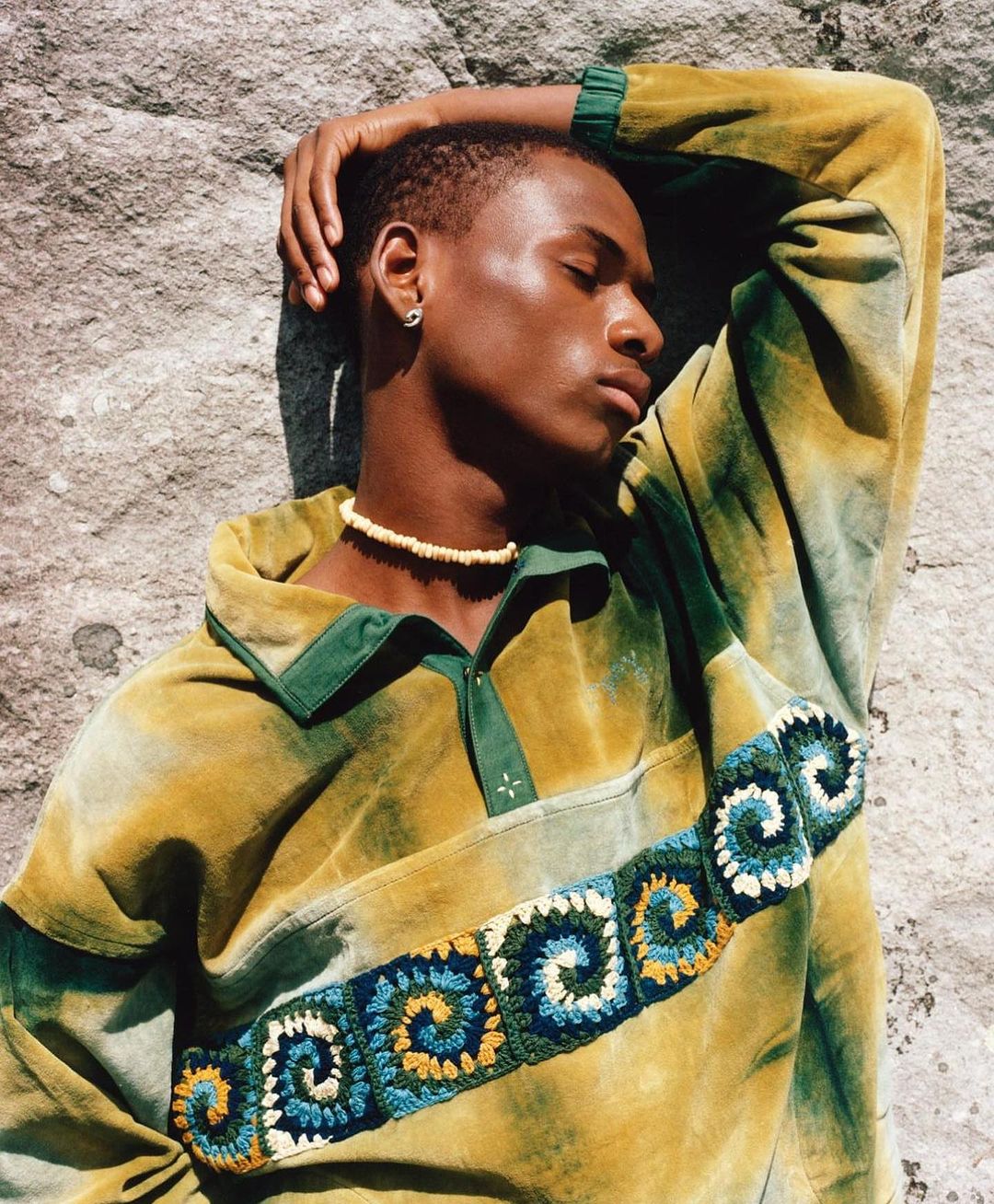 Adult male fashion model poses outdoors in front of a rock wall while wearing a tie-dye long sleeve shirt with a beaded necklace.