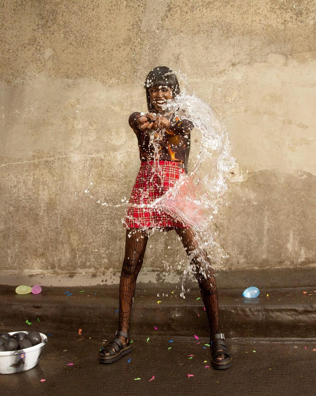 An adult female smiling and splashing water with water balloons in an outdoor setting with a concrete backdrop, wearing a red plaid skirt.