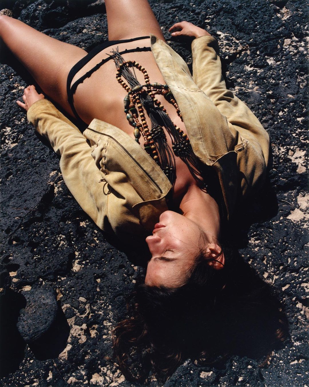 A woman in a jacket and black underwear lies on dark, rough volcanic rock in an outdoor setting, wearing a beaded necklace.