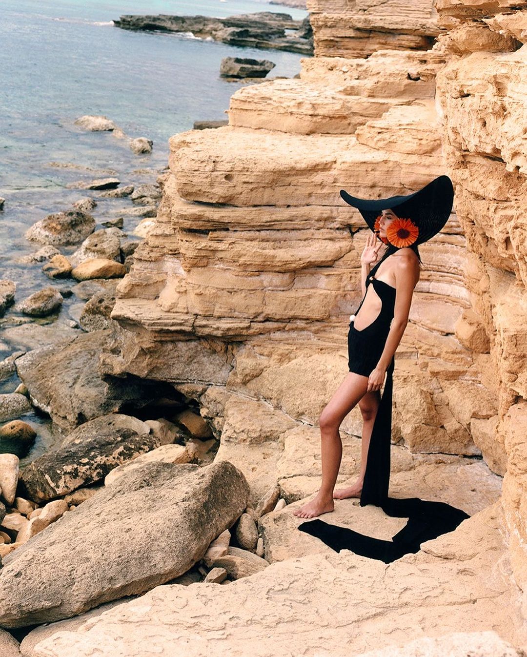 Woman in black swimsuit and wide-brimmed hat posing on rocky beach with ocean background in a fashion editorial.