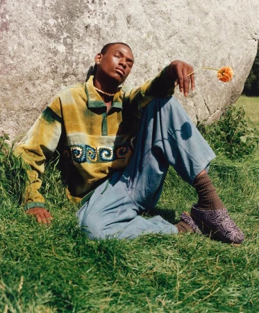Black man seated on grassy ground leaning against a large boulder holding a small orange flower, wearing patterned shirt and blue trousers.