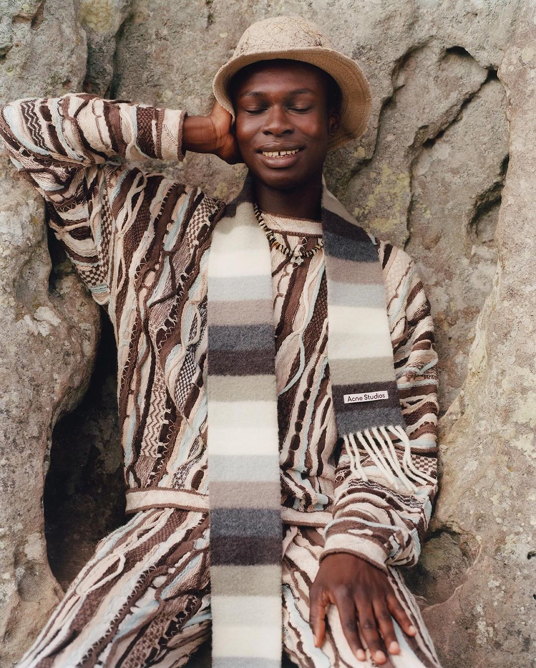 Stylish adult man poses in a textured sweater, pants, and hat, accented by a striped scarf against a rustic rock backdrop.