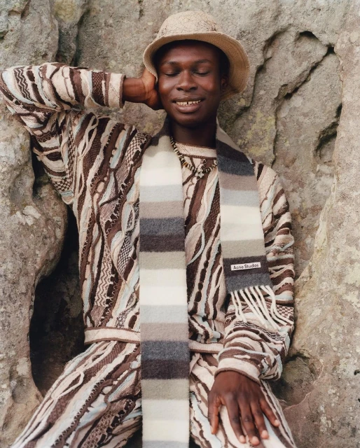 Stylish adult man poses in a textured sweater, pants, and hat, accented by a striped scarf against a rustic rock backdrop.
