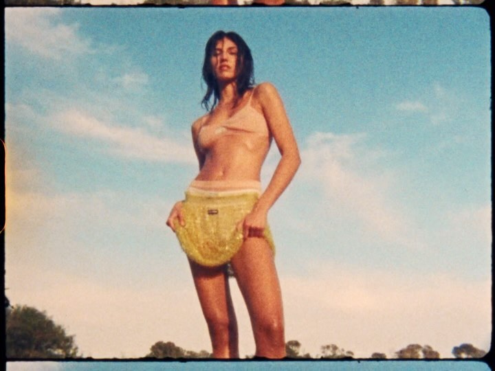 Woman poses in yellow skirt and light peach top against blue sky and neutral background, captured in a vintage aesthetic.