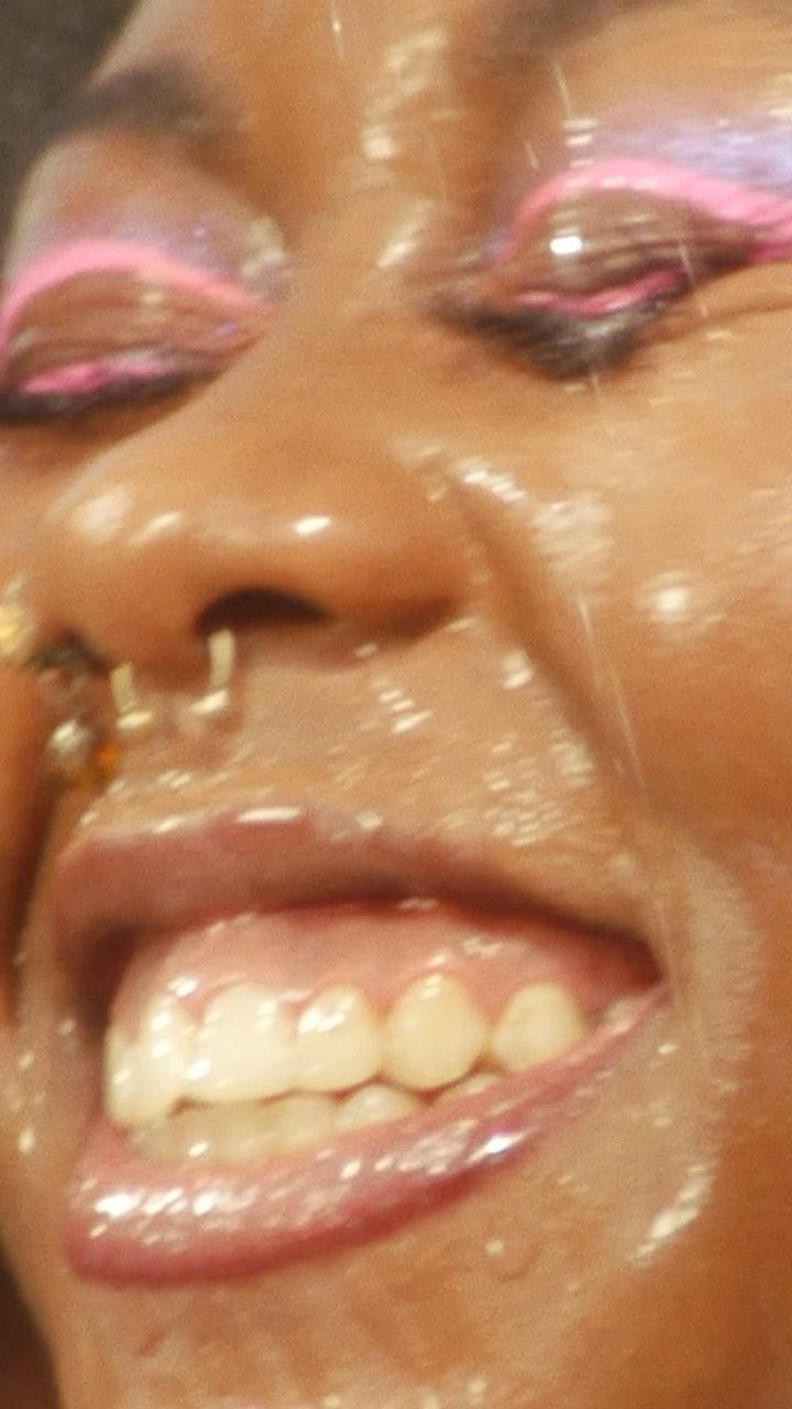 Close-up shot of an African American woman's face, featuring bright pink and purple eye makeup, nose piercings, and glossy lips.