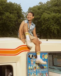 Young man smiling while sitting on top of an ice cream van wearing a sleeveless denim vest and white shoes.