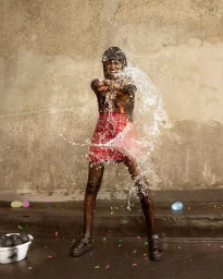 Black woman splashing water towards camera wearing plaid skirt and dark patterned top, by photographer Kaj Jefferies
