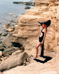 Model in wide black sun hat with orange flower and black cut-out dress standing on coastal rocks by the ocean.